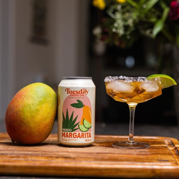A mango, a "Tuesday Cocktail Club Mango Margarita" can, and a margarita in a glass with a lime.