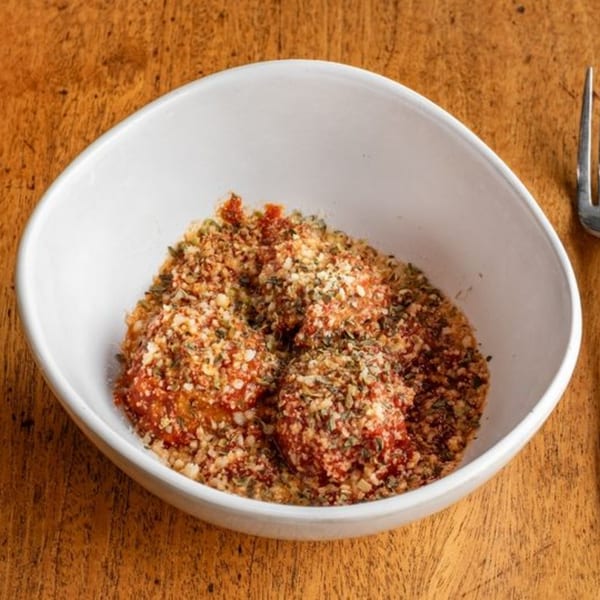 Three meatballs covered in red sauce and breadcrumbs in a white bowl on a wooden table.