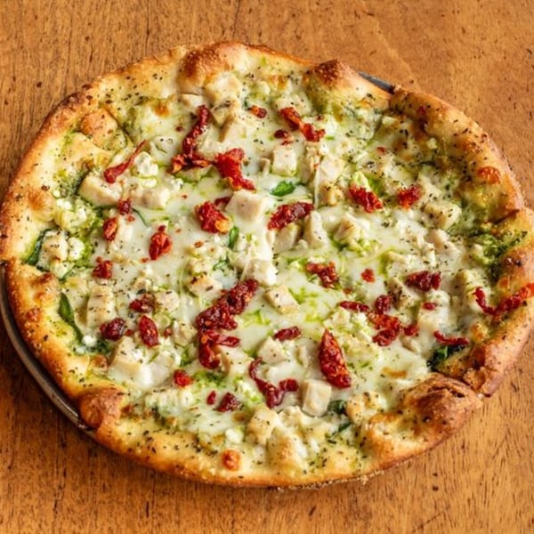Little Pizza Paradise's pesto pizza with chicken, sun-dried tomatoes, and feta cheese on a wooden table.