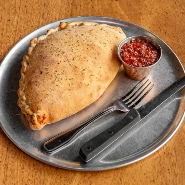 A golden-brown calzone with a side of marinara sauce, fork, and knife on a metal plate.
