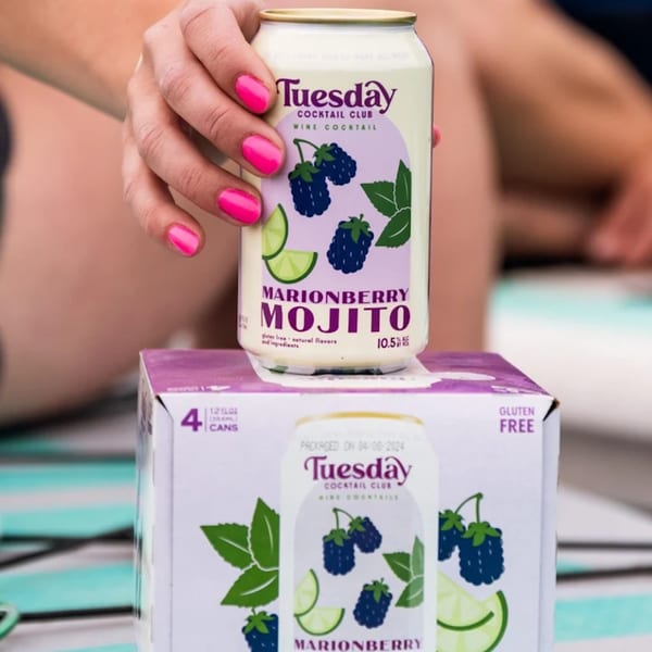 A hand with pink nails holds a Marionberry Mojito can above a 4-pack box.