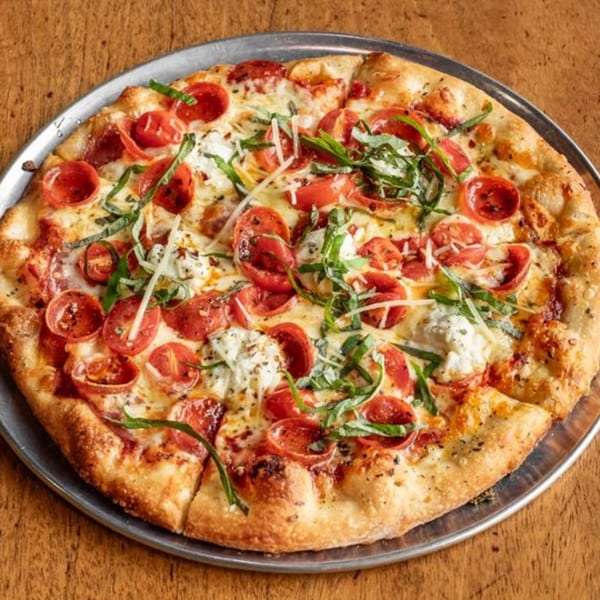 A Little Pizza Paradise pizza with pepperoni, tomatoes, fresh basil, and cheese on a metal pan.